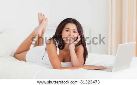 cute woman lying on bed with crossed legs and laptop looking into camera in bedroom
