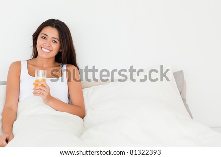 smiling woman holding orange juice sitting in bed looking into camera in bedroom