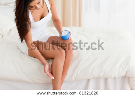 smiling woman putting creme on her legs while sitting on bed in bedroom