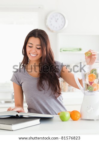 Attractive woman consulting a notebook while filling a blender with fruits in the kitchen