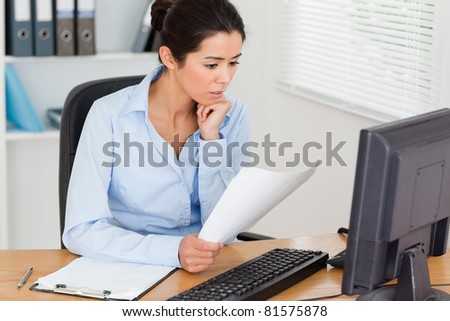 Gorgeous woman looking at a computer screen while holding a sheet of paper at the office