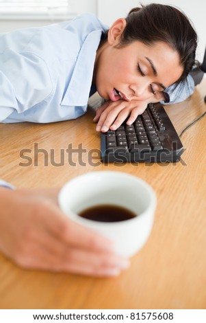 Gorgeous woman sleeping on a keyboard while holding a cup of coffee at the office