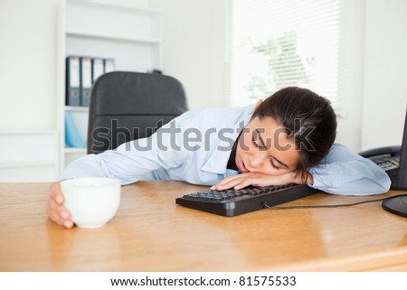 Frontal view of a pretty woman sleeping on a keyboard while holding a cup of coffee at the office