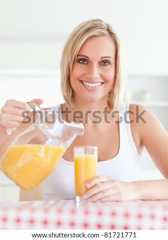 Close up of a smiling woman sitting at a kitchen table filling orange juice in a glass smiling into the camera