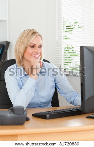 Smiling blonde woman with chin on hand behind a desk looking at screen in an office