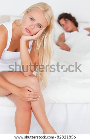 Portrait of an unhappy woman sitting on a bed while her fiance is sleeping with the camera focus on the foreground