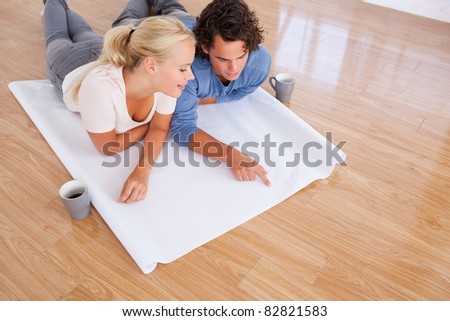 Cute man showing a point on a plan to his fiance while lying on the floor Cute man showing a point on a plan to his fiance while lying on the floor