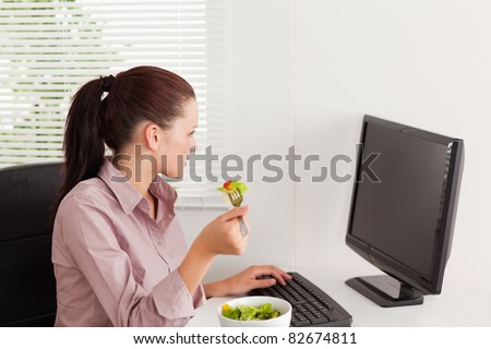 A businesswoman is eating salad whilst typing and looking to screen of her pc