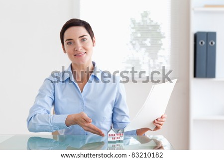 Short-haired Woman holding papers pointing at a model house in an office