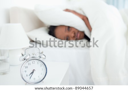 Exhausted woman covering her ears while her alarm clock is ringing in her bedroom