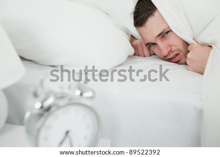 Annoyed man covering his ears while his alarm clock is ringing in his bedroom