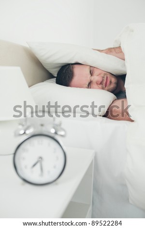 Portrait of a unhappy man covering his ears while his alarm clock is ringing in his bedroom