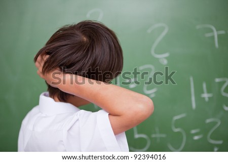Schoolboy thinking while scratching the back of his head in front of a chalkboard