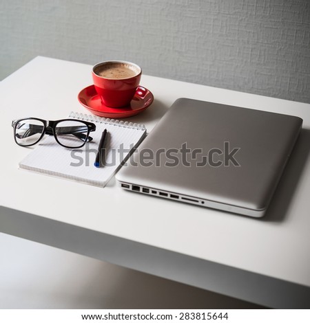 White office table on which laptop, red cup of coffee, glasses, notebook and pen