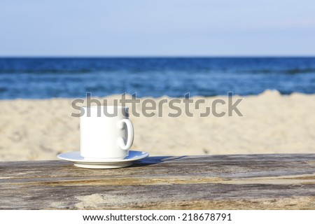 White mug stands on a wooden table which stands on the sand beach at the Baltic Sea coast