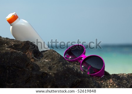Pink sunglasses and bottle of sun-protective lotion on a rock with blurred sea in background
