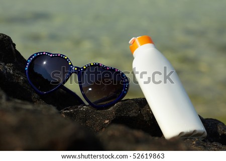 Dark blue heart shaped sunglasses and bottle of sun protecting lotion on a rock with blurred sea in background