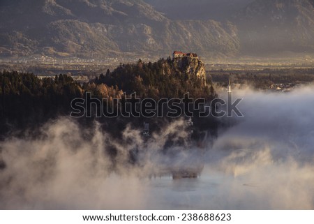 Mystical Sunrise over Lake in the Mountains. Mist is over the lake covering the church almost in full. Castle and surrounding Mountains are basking in the sun. Scenic and atmospheric sunrise on Bled.
