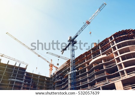High building under construction. Side with cranes against blue sky with sun glare.