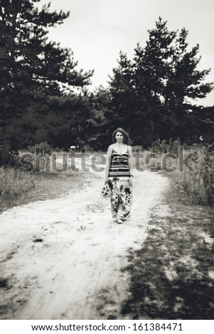 Black and white photo of girl in long dress walking on road in field