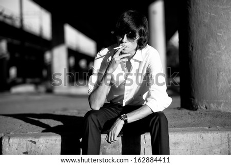Black and white portrait of man in white shirt smoking cigarette against city landscape