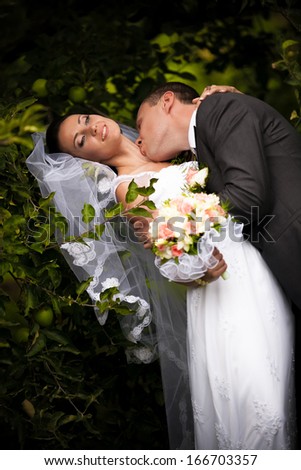 Handsome groom kissing passionately bride in neck neck under tree