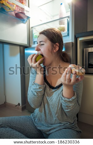 Closeup portrait of woman in pajamas biting apple at kitchen at night
