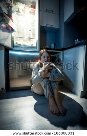 Photo of young woman eating next to refrigerator at night
