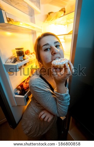 Closeup portrait of woman biting donut at night near open fridge