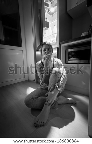 Black and white photo of woman sitting next to fridge at night