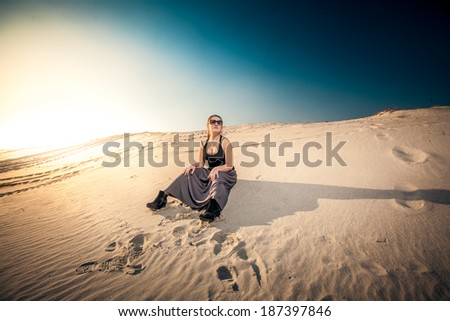 Beautiful woman in long skirt sitting on sand dune against blue sky and sun