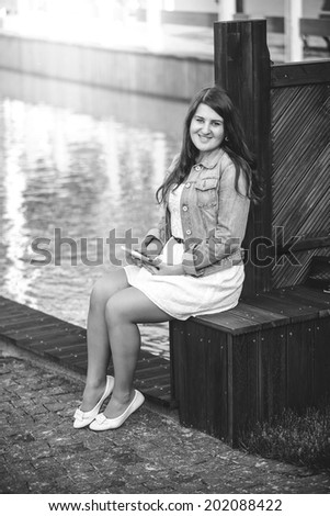 Black and white photo of young woman sitting on bench near river with tablet