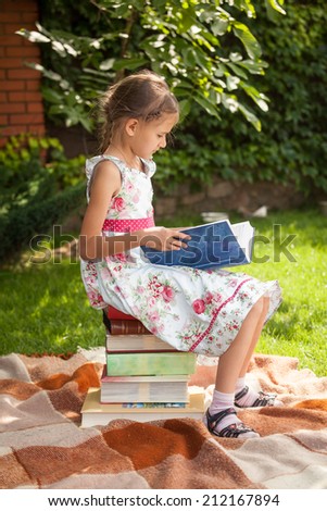 Little girl in cute dress sitting on pile of books and reading