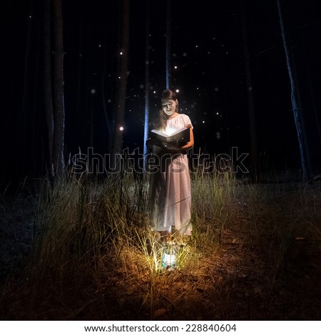Beautiful woman in long dress reading big old book at mysterious forest with fireflies