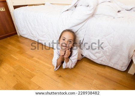Smiling little girl lying under bed at home and looking at camera