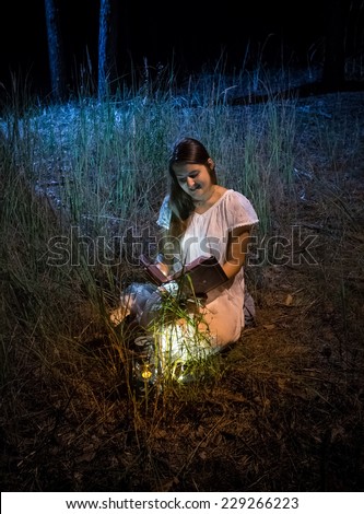 Young woman sitting at night forest with lantern reading big old book