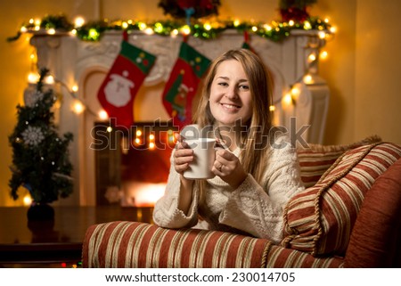 Cute young woman sitting at fireplace with cup of tea at Christmas