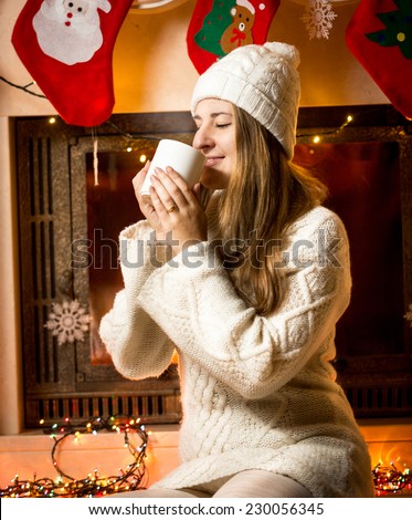 Portrait of cute woman in knitted sweater drinking hot tea next to fireplace