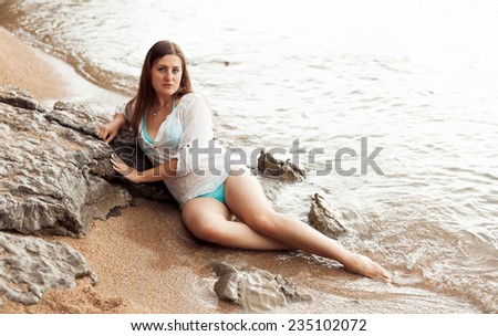 Sexy brunette model in wet shirt lying on rock at sea beach