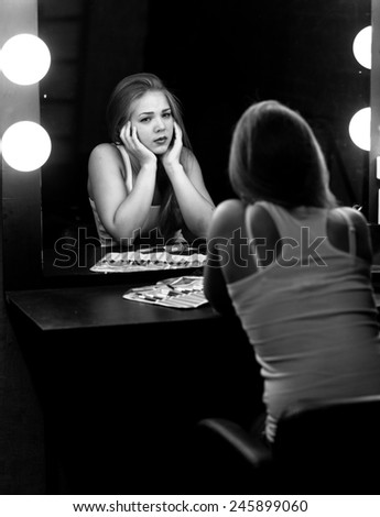 Black and white portrait of sad woman looking at reflection in mirror with bulbs