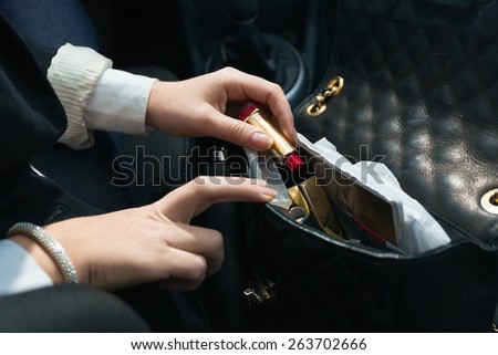Young woman sitting in car and taking lipstick out of handbag