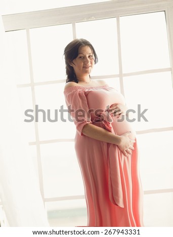 Toned photo of pregnant brunette woman posing against big window