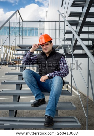 Male architect wearing red hard hat sitting on metal staircase at factory