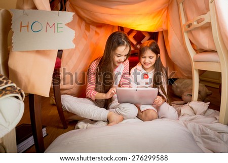 Happy girls sitting in house made of blankets and using digital tablet