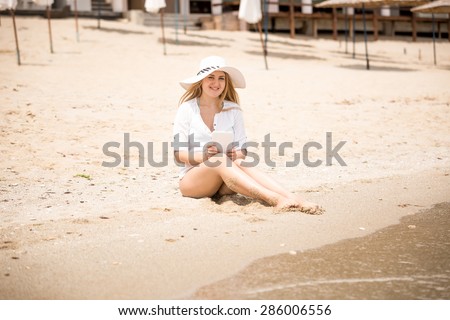 Beautiful young woman in hat sitting on beach and using digital tablet
