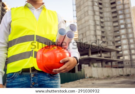 Closeup toned photo of foreman holding helmet and blueprints against building site