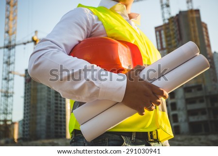 Closeup photo of man in shirt and jacket holding hardhat and blueprints on building site