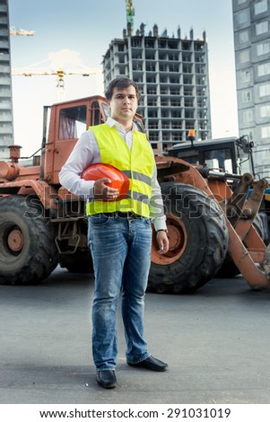 Foreman in yellow safety jacket posing next to bulldozer on building site