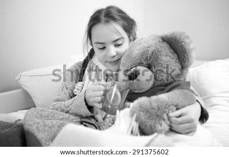 Black and white portrait of cute girl lying in bed and giving pills to teddy bear