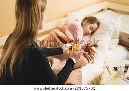 Young caring mother giving hot tea to daughter lying in bed